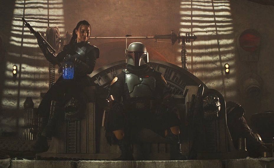 Promo pic for The Book of Boba Fett. Boba is sitting in a majestic chair surrounded by weapons. Fennec Shand is leaning against the chair.